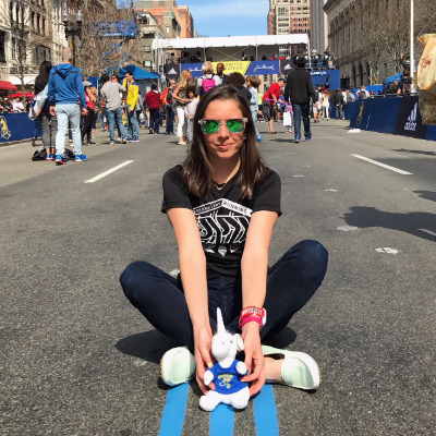 A female wearing sunglasses sitting with a toy stuffed unicorn near the Boston Marathon finish line