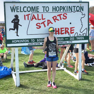 A female runner standing in front of a sign about the Boston Marathon