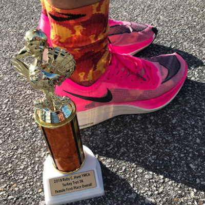 A photo of a first place 5K turkey trot trophy with a person wearing pink Nike VaporFly ZoomX Next % running shoes behind it